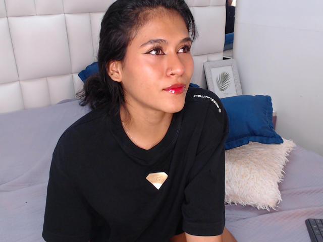 AnnieDarling with dark hair, brown eyes, and red lipstick, wearing a black shirt with a diamond-shaped emblem, sitting on a bed with blue and white pillows.