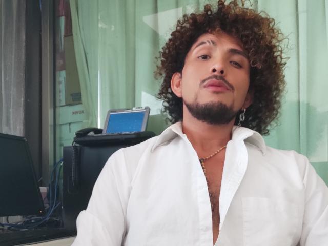 Dandann with curly brown hair, beard, and mustache, wearing a white button-down shirt, necklace, and earring, looking slightly bored. Background includes office equipment.