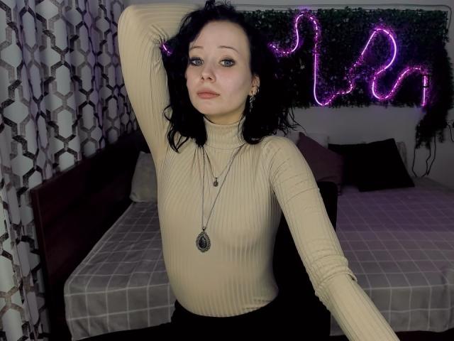 EliseVance with black curly hair, pale skin, and blue eyes, wearing a beige ribbed turtleneck, black skirt, and necklace, arm raised, looking slightly annoyed, purple neon butterfly in background.