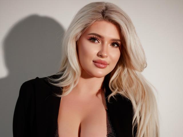 CocoSecrets69 has blonde hair, full lips, and large tits. She's wearing a black top with a deep cleavage. Her long hair and shadow are visible.