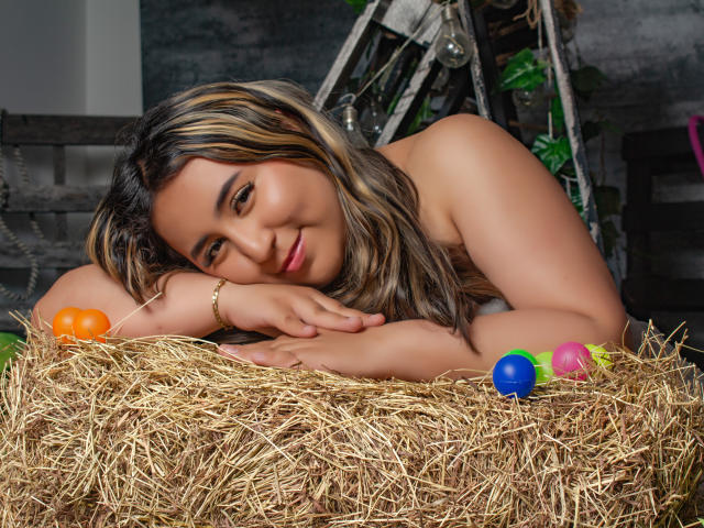 SalomettX with medium brown skin, blonde and black hair, lying on hay, head on arm, smiling, colorful eggs around, bracelet on wrist, soft light, relaxed, sexy.