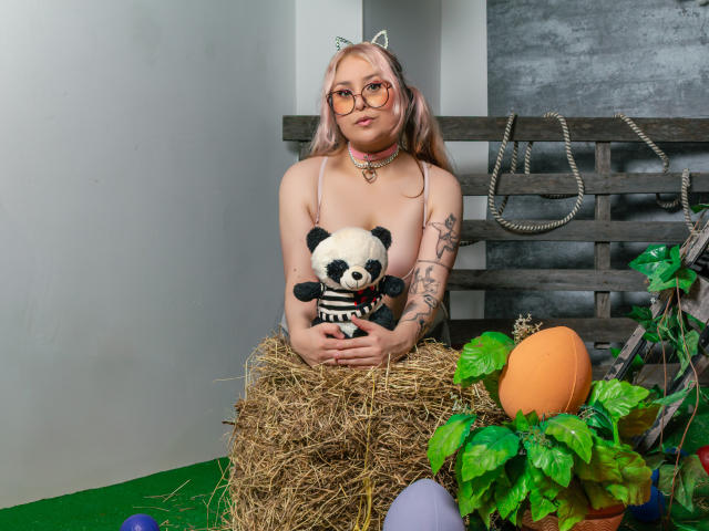 DandelionQueen with pink hair, glasses, cat ears, choker, tattoos, holding panda stuffed toy, sitting on hay, orange pumpkin, green plant, wooden wall, and rope in background.