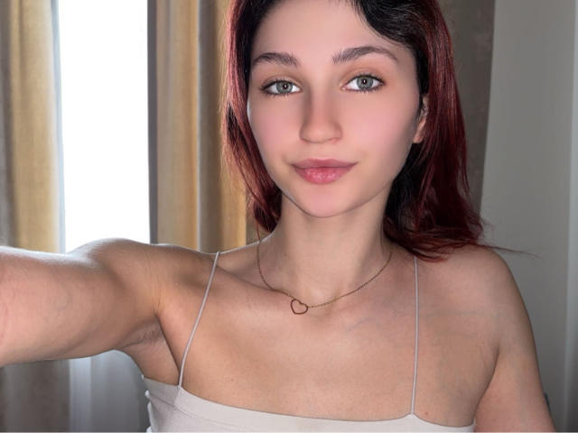 CarmelinaLove has pale skin, blue eyes, and red hair, wearing a white spaghetti-strap top, showing her small tits, and a delicate necklace with a small ring.