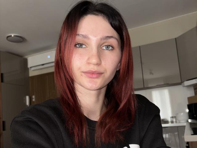 CarmelinaLove with red-brown hair, green eyes, and light skin, wearing a black shirt, has a small zit on her nose and a subtle smile.