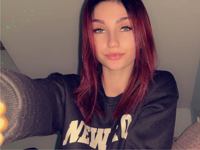 CarmelinaLove has red hair, green eyes, and a cute face with a slight smile. She's wearing a black New York sweatshirt and grey socks.