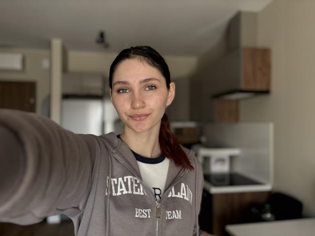 CarmelinaLove with a smug smile, brown eyes, and a red ponytail, wearing a gray hoodie, taking a selfie with her right arm extended. Boobs are not visible.