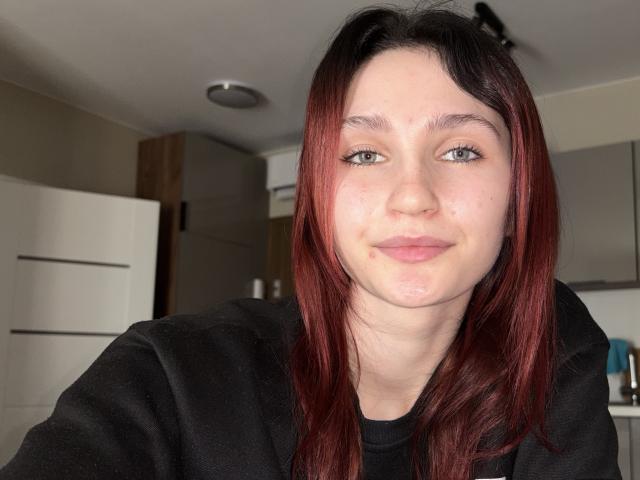 CarmelinaLove has pale skin, green eyes, and red-brown hair in a black hoodie, with a subtle smile and slight freckles on her face. Her boobs are not visible.