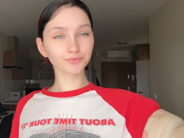 CarmelinaLove, with pale skin and green eyes, has dark brown hair in a ponytail, wearing a red and white rag tee, and a fluffy beige sleeve, smiling softly.