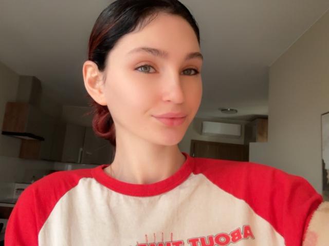 CarmelinaLove with pale skin, green eyes, and black hair in a red and white raglan shirt, smiling slightly, showing her cute nose and subtle pink lipstick.
