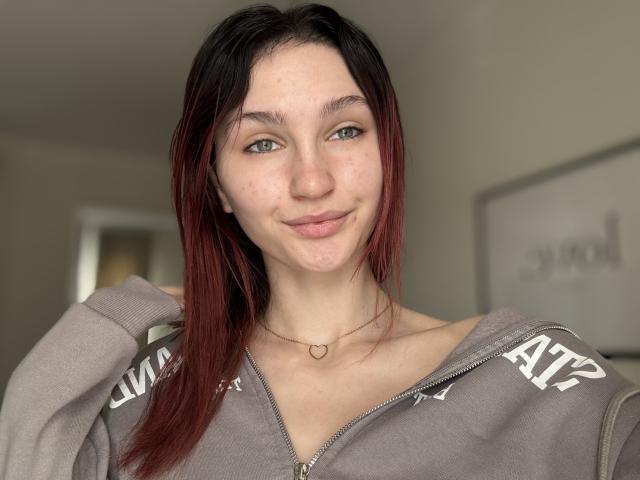 CarmelinaLove with pale skin, green eyes, and red-dyed black hair, smiling slightly, wearing a gray zip-up hoodie, and a heart necklace. Her tits are small and unvisible.