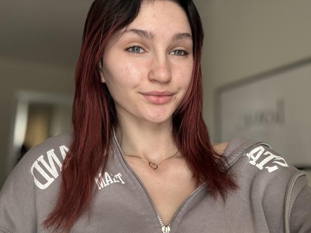 CarmelinaLove has light skin, green eyes, and red-brown straight hair. She's wearing a gray zip-up hoodie with white letters, and a small heart necklace. She's smiling softly.