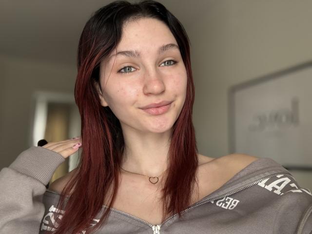 CarmelinaLove with red and black hair, blue eyes, freckles, and a small heart necklace, smiling gently in a gray off-shoulder zip-up, hand near her head.