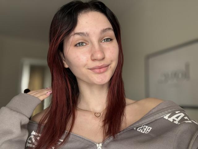 CarmelinaLove has pale skin, green eyes, and red-black dyed hair, wearing a grey off-shoulder zip-up, heart necklace, and a black ring, smiling softly.
