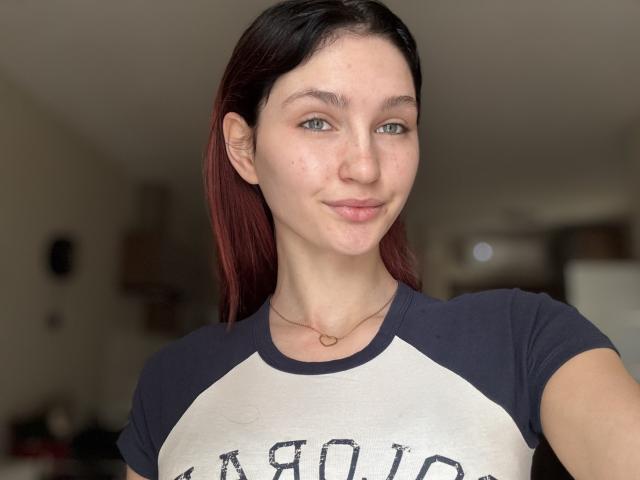 CarmelinaLove, with light skin and green eyes, has dark red hair, wearing a black and white raglan shirt, and a small heart necklace, smiling softly at the camera.