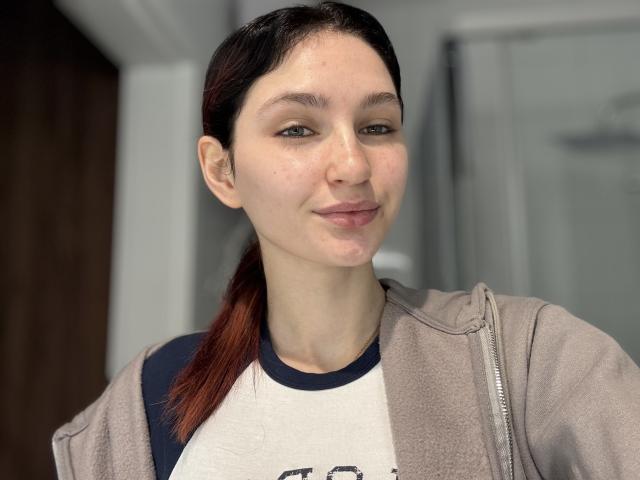 CarmelinaLove with pale skin, dark brown ponytail, subtle smile, wearing a beige jacket over a navy and white shirt, slightly shiny face, looking at the camera.