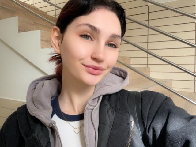 CarmelinaLove with fair skin, black hair, and green eyes, wearing a gray hoodie and black jacket, smiling with pink lips, and a gold necklace. Stairs in the background.