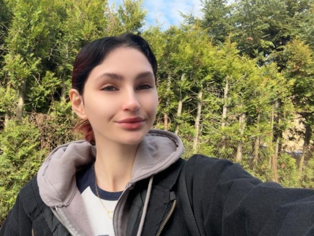 CarmelinaLove has light skin, dark brown hair in a ponytail, small nose, and subtle smile. She's wearing a black jacket with a gray hoodie. Her tits are small.