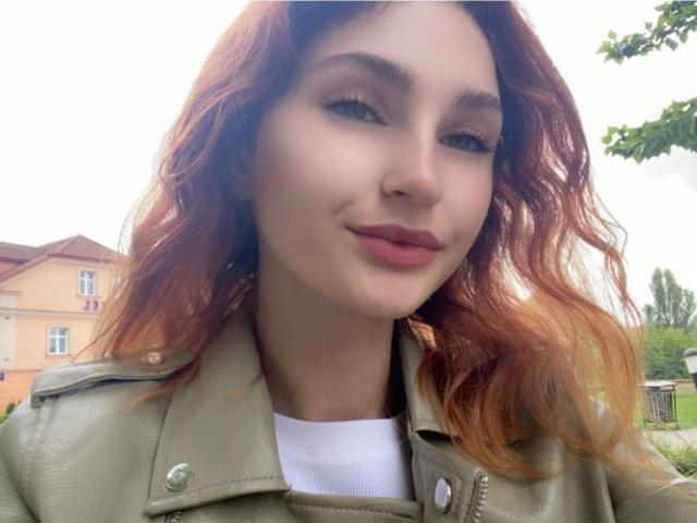 CarmelinaLove with wavy red hair, green jacket, and white shirt, has a cute smile and full pink lips. She's looking at the camera with a slight smirk.
