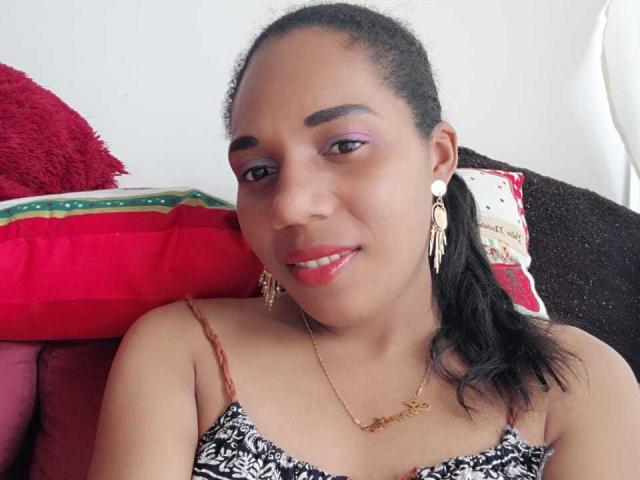 CattyMoon with dark skin, purple eyeshadow, red lipstick, black hair in ponytail, wearing patterned top, gold necklace, earrings, smiling, sitting on red and white pillows.
