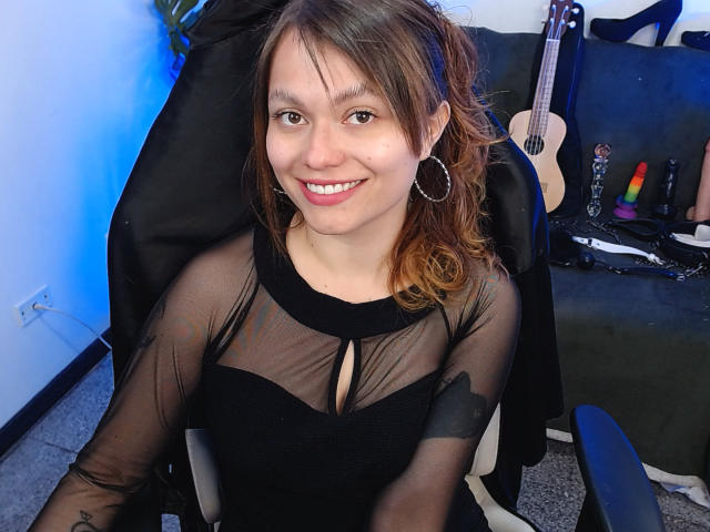 Smiling BlackBerries with brown hair, hoop earrings, and sheer black top, showing small boobs, sitting in chair, with a guitar, dildo, and vibrator in the background.