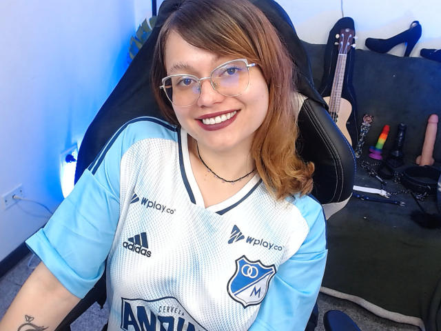 BlackBerries with light brown hair, glasses, smiling, wearing a blue and white sports jersey, sitting in a chair, with a guitar and sex toys in the background.