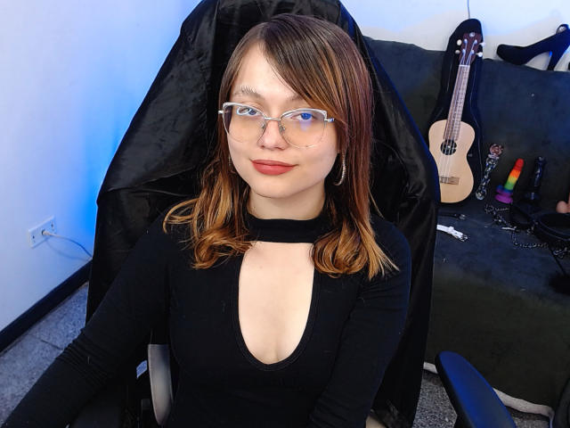 BlackBerries with glasses, brown hair, black choker, and black top, showing cleavage, sitting in chair, black jacket behind, guitar and sex toys on green sofa.