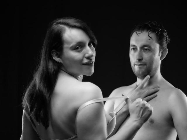 JeremmyAndStacy with tits exposed, holding a strap, smiling at JeremmyAndStacy's chest, who's shirtless with a finger on his chin, in a black-and-white photo.