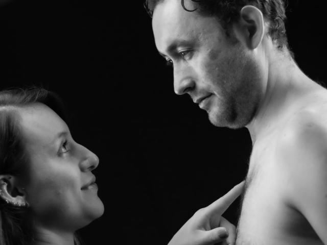 Zwart-wit foto van Jeremmy met een kaal torso, die naar het profiel van Stacy staart, haar hand raakt zachtjes zijn borst, beiden met een serieuze uitdrukking, donkere achtergrond..