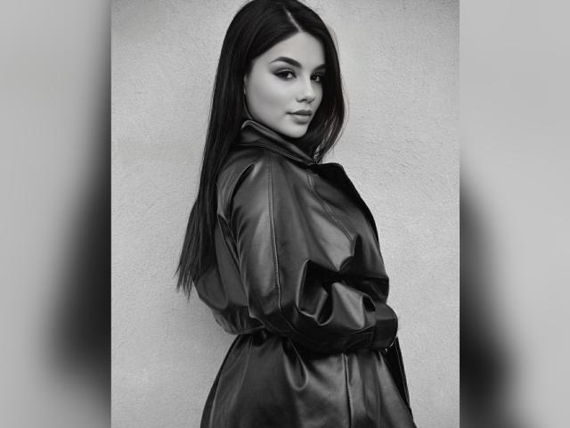 VioletX69 with long dark hair, wearing a shiny black leather dress, looking over her shoulder, hands in pockets, grey background, black-and-white photo, sexy, confident, cute.