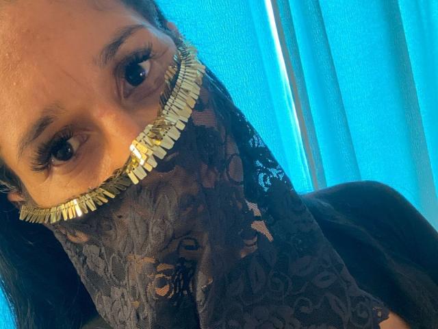 Le visage de KiraTaleb, portant un masque en dentelle noire avec des bords dorés, laissant apparaître ses yeux et ses longs cils, sur un fond bleu. Elle regarde l'appareil photo..