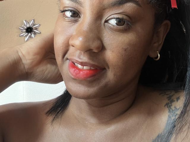 TiffanySammy's face, dark brown skin, red lipstick, small gold hoop earring, black hair, hand behind head, tattoo on right shoulder, brown eyes, subtle smile.