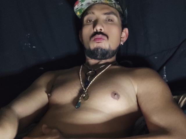 Shirtless Dandann with a mustache, bearded, wearing a patterned cap, necklace with a green stone, and earrings, lying down, dark background, looking at camera.