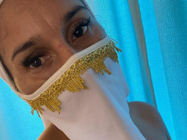 Il volto di KiraTaleb, con occhi scuri, indossa una maschera bianca con bordi dorati, ornati di gioielli e frange, e un velo bianco che copre la testa, mostrando solo gli occhi e la maschera in una foto illuminata di blu..