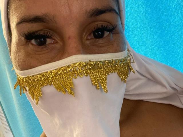 Kira Taleb, avec une peau brun clair, des yeux foncés et de longs cils, portant un masque blanc orné de sequins dorés, qui couvre partiellement sa bouche et son nez. Fond bleu..