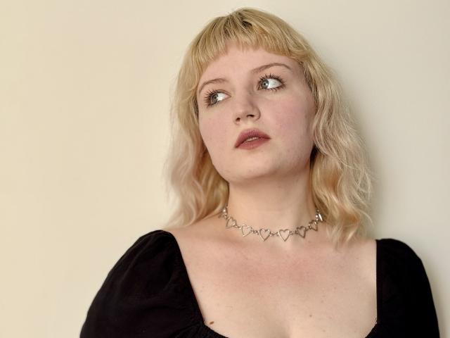 TeresaLibson ha i capelli biondi e ondulati, la pelle chiara e gli occhi azzurri, indossa una maglia nera con maniche a sbuffo e una collana d'argento a forma di cuore. Il suo seno è leggermente visibile..