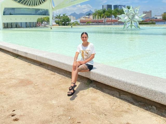 SamanthaBss, with black hair in a bun, white t-shirt, black shorts, and sandals, sits on a stone edge by a clear blue water fountain. Visible tattoo on her left leg.