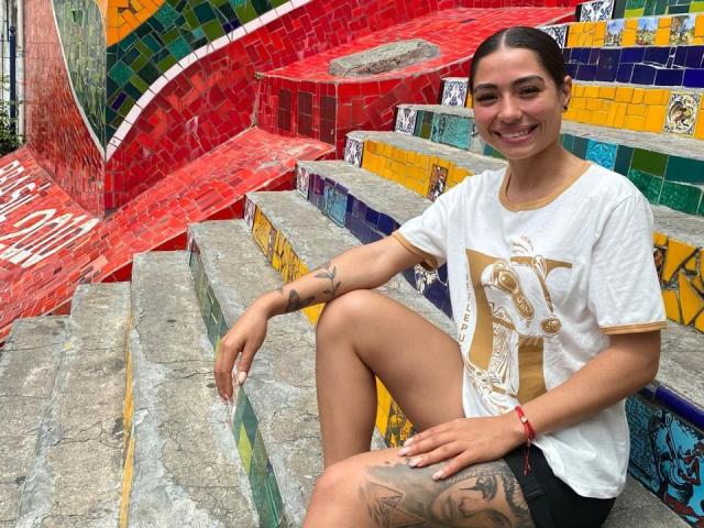 SamanthaBss with tattoos, smiling, wearing a white tee and black shorts, sitting on colorful tiled stairs, has a red bracelet and brown hair in a ponytail.