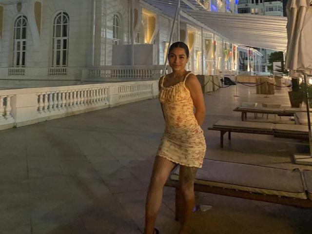 SamanthaBss with tan skin, brown hair in a ponytail, wearing a floral, short, sleeveless dress, standing with hands behind her back, smiling at night in an outdoor setting.