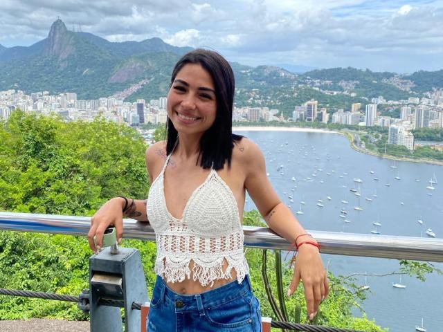 SamanthaBss with black hair, white crochet top, and blue jeans, smiling, leaning on a metal railing, showing her small tits, with a city and water background.