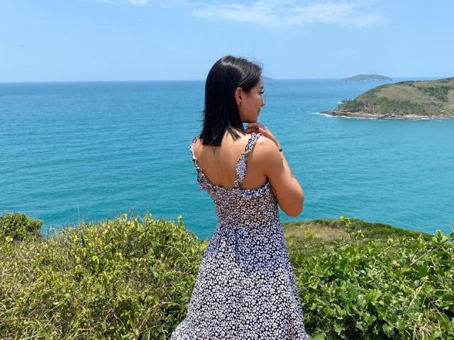SamanthaBss in a floral dress, back to camera, black hair, blue ocean, green shrubs, sunny, standing on cliff, looking right, cute ass, small tits, summer vibe.