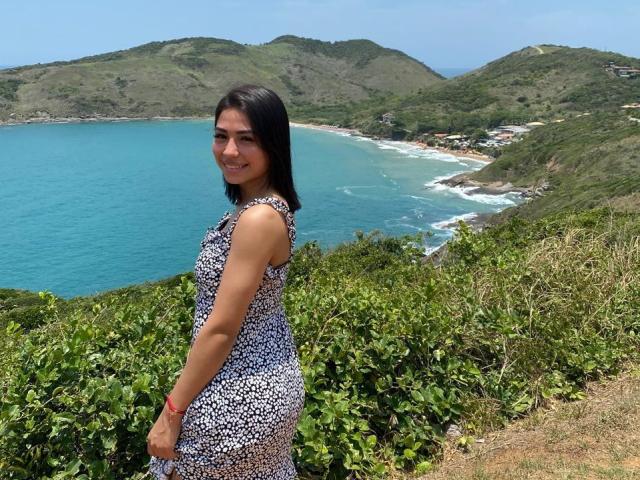 SamanthaBss with black hair, wearing a sleeveless white and blue floral dress, smiling at the camera, standing on a grassy hill with a blue ocean and green hills.