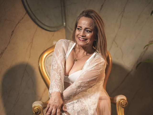 MaryMidori with medium tits, smiling, wearing a white lace robe, exposing her cleavage, sitting on a golden chair, brown hair, green eyes, and a cross necklace.
