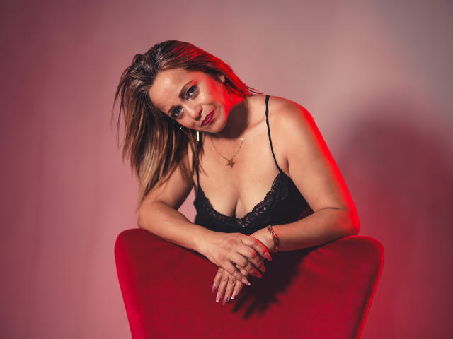 MaryMidori with red highlights in her brown hair, wearing a black lace top, leaning on a red chair, hands together, smiling slightly, red light on her right side.
