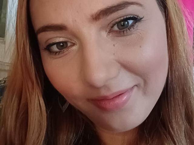 NicoleBisous has light brown eyes, subtle makeup, and a slight smile with pink lips. She has blonde hair, a small nose, and a few tiny moles on her face.