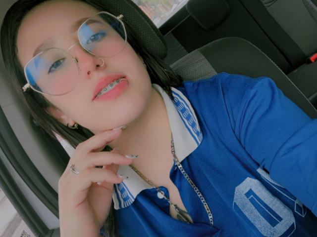 NatalieHarris with glasses, blue shirt, white collar, black hair, pink lips, metal chain, ring, and pierced tongue, sitting in car, hand near face, smiling slightly.