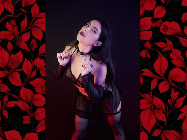 SamantaKi has black hair, a choker, and wears a sheer black dress, exposing her tits and tattoos, with red floral-patterned sides, leaning forward with a seductive look.