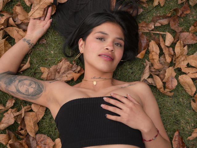 AlaiaStone, with black hair, lies on grass, surrounded by dry leaves, wearing a black tube top, showing tattoos, and a necklace, with hands on her chest, smiling.