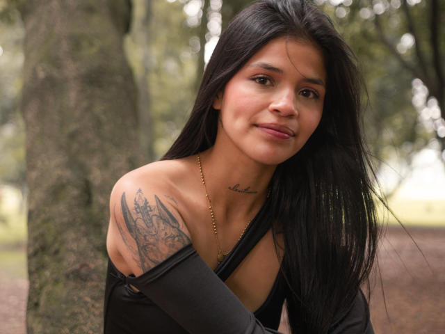 AlaiaStone mit langen, schwarzen Haaren, mittlerer Brustgröße, tätowierter Schulter, einem schwarzen Top mit einem Schulterauschnitt, einer goldenen Halskette und einem leichten Lächeln, steht neben einem Baum in einem Wald..