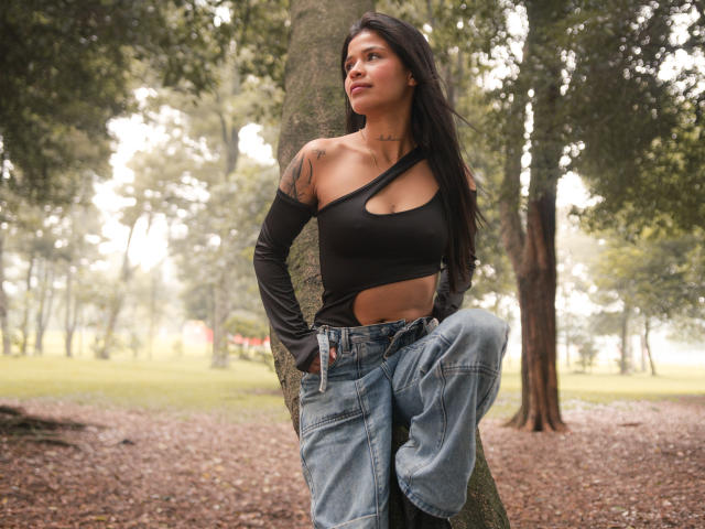 AlaiaStone with long black hair, wearing a black one-shoulder top showing her tits and cut-out midriff, and blue jeans, standing against a tree in a forest.