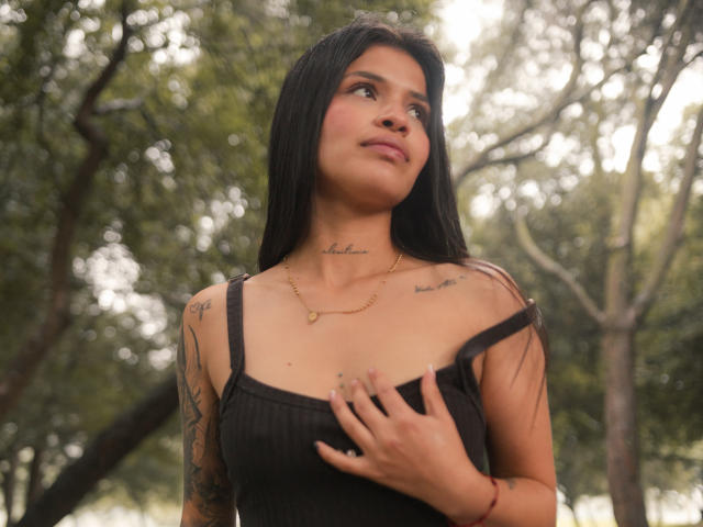 AlaiaStone with long black hair, small tits, tattoos, black ribbed tank top, gold necklace, looking up, right hand on chest, forest background, soft light, slight smile.